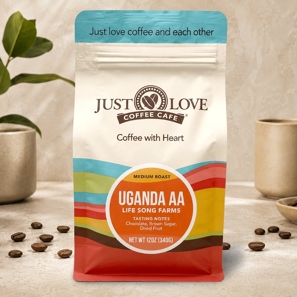 A 12 oz bag of Just Love Coffee Cafe Uganda AA medium roast coffee stands on a light stone surface with scattered coffee beans. The bag is cream-colored with a blue top band, multicolor stripes, and a large orange circular label.